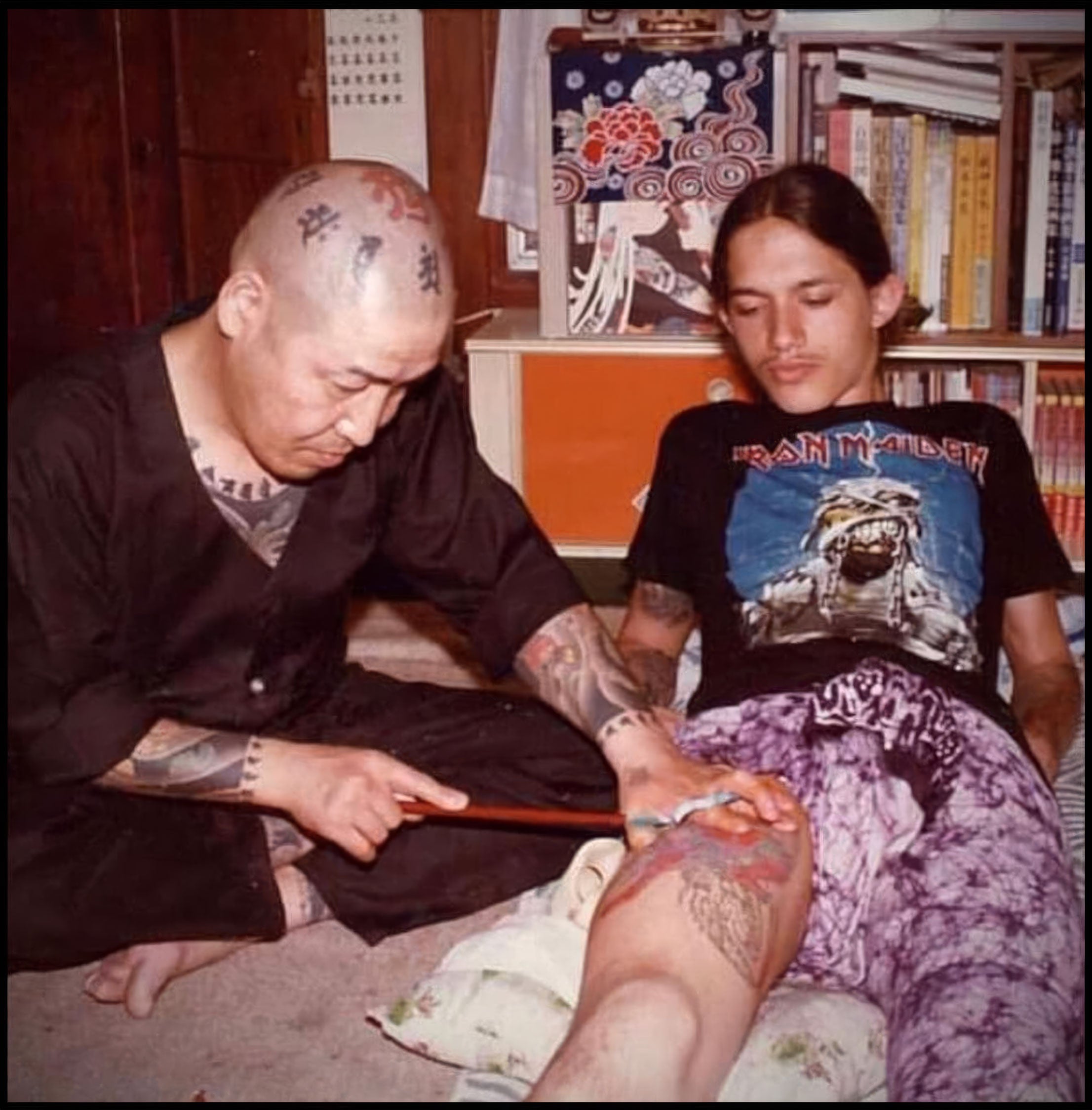 A photo from the 80s, a bald monk with a tattoo head. hand tattooing a teenager with an iron maiden shirt