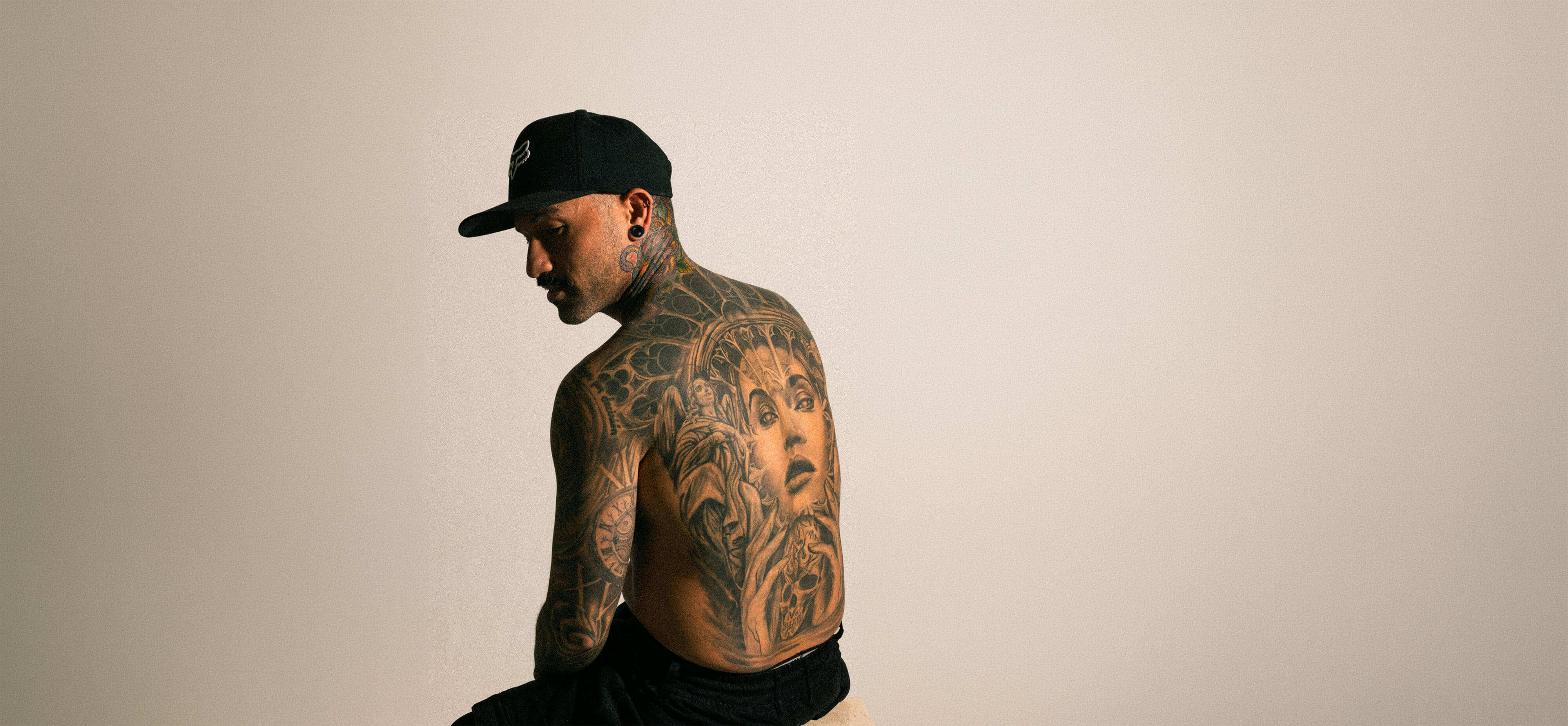 man shirtless in a hat sitting in a chair, moody lighting, with a big back tattoo