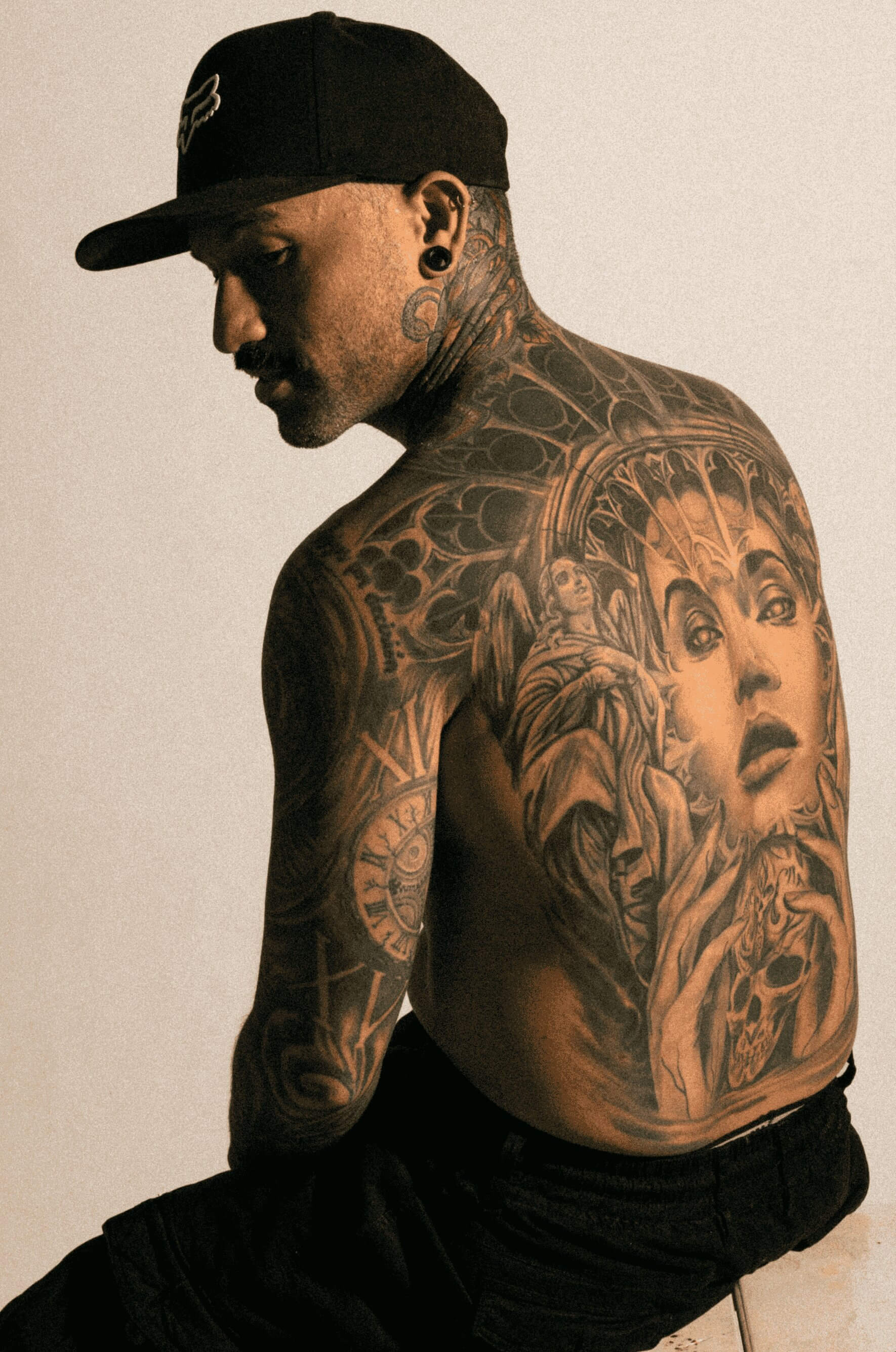 man shirtless in a hat sitting in a chair, moody lighting, with a big back tattoo